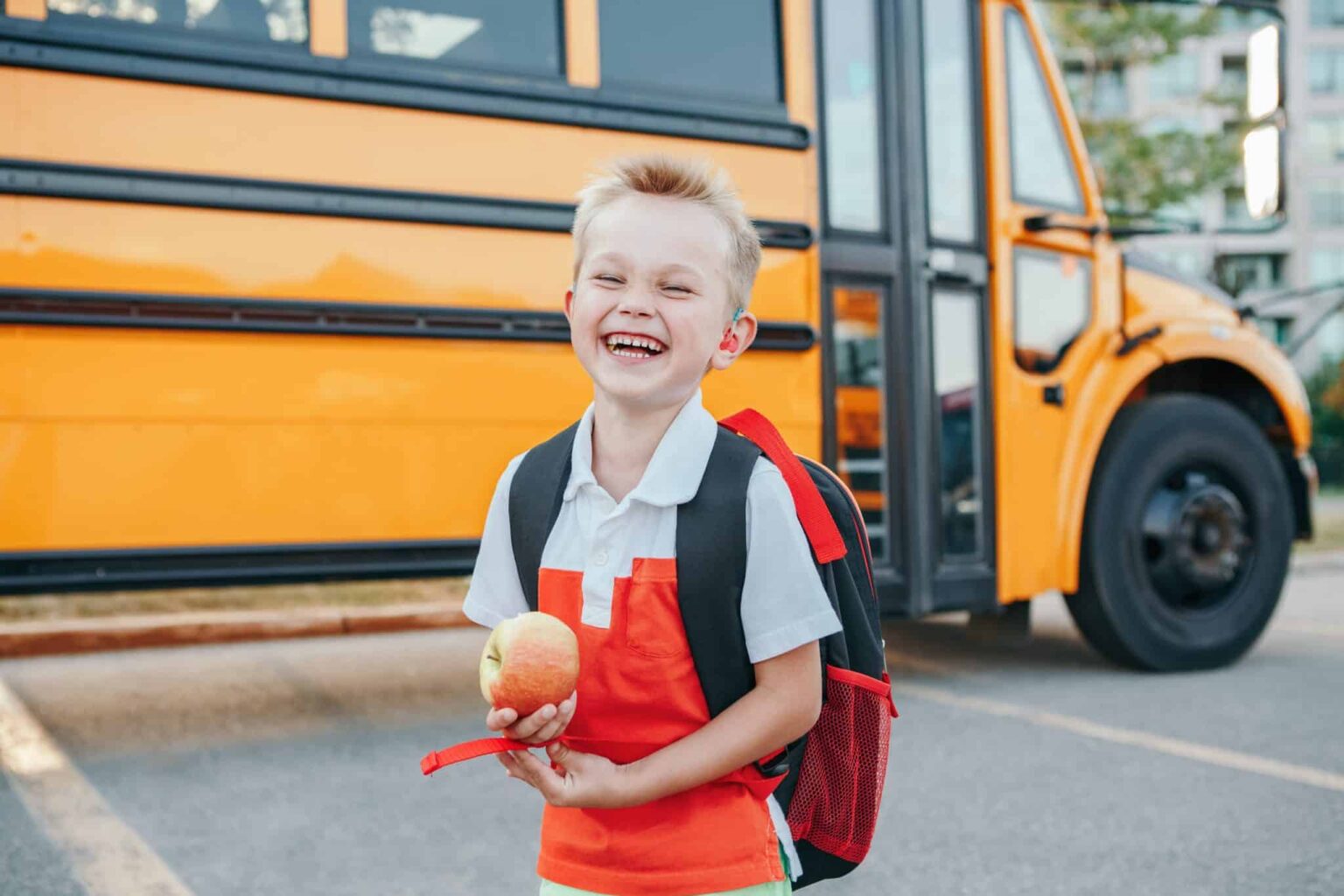Child wearing pediatric hearing aid Smiling child wearing a backpack and holding an apple. There is a schoolbus parked behind them.